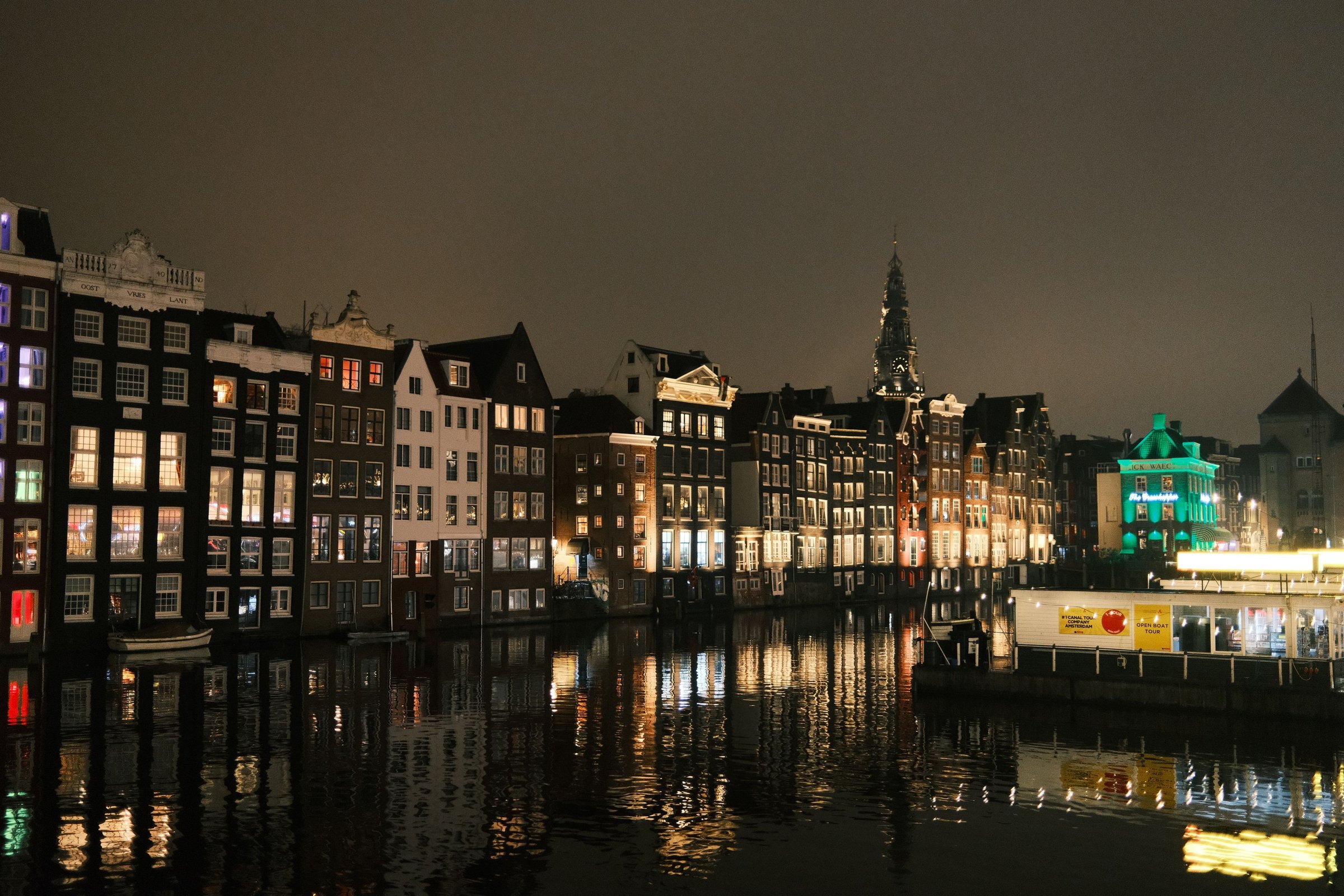 Deretan rumah kanal Amsterdam menyala di malam hari dengan pantulan cahaya di air.
