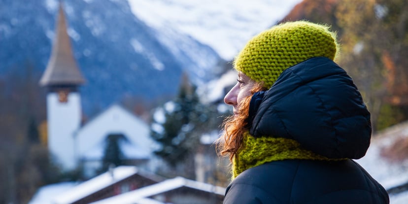 Seorang perempuan berjaket hitam dan beanie hijau memandang gereja dan desa Lauterbrunnen yang bersalju, pegunungan Alpen di kejauhan.