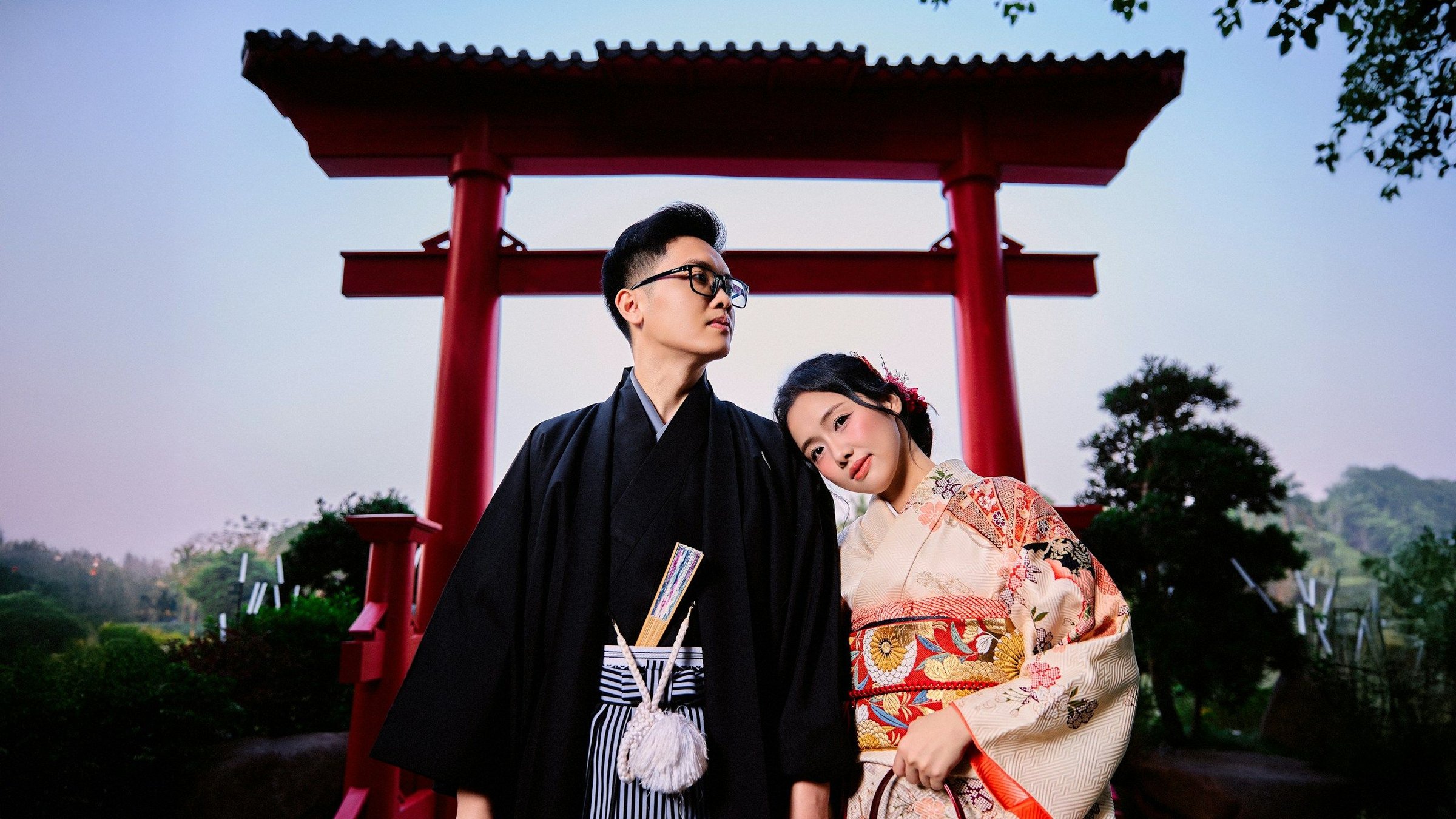 Pasangan berbusana kimono tradisional berdiri di depan gerbang torii merah saat senja