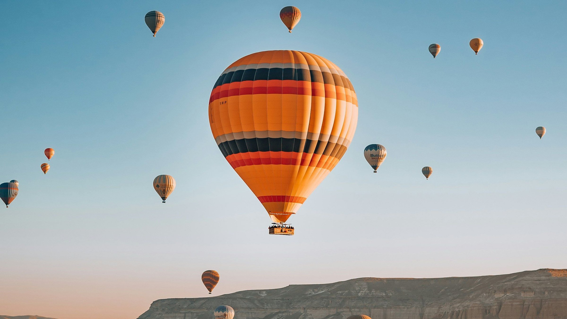 Puluhan balon udara warna-warni mengambang di atas cerobong peri Cappadocia saat matahari terbit, dengan satu balon oranye besar di tengah dan lanskap batuan Turkiye