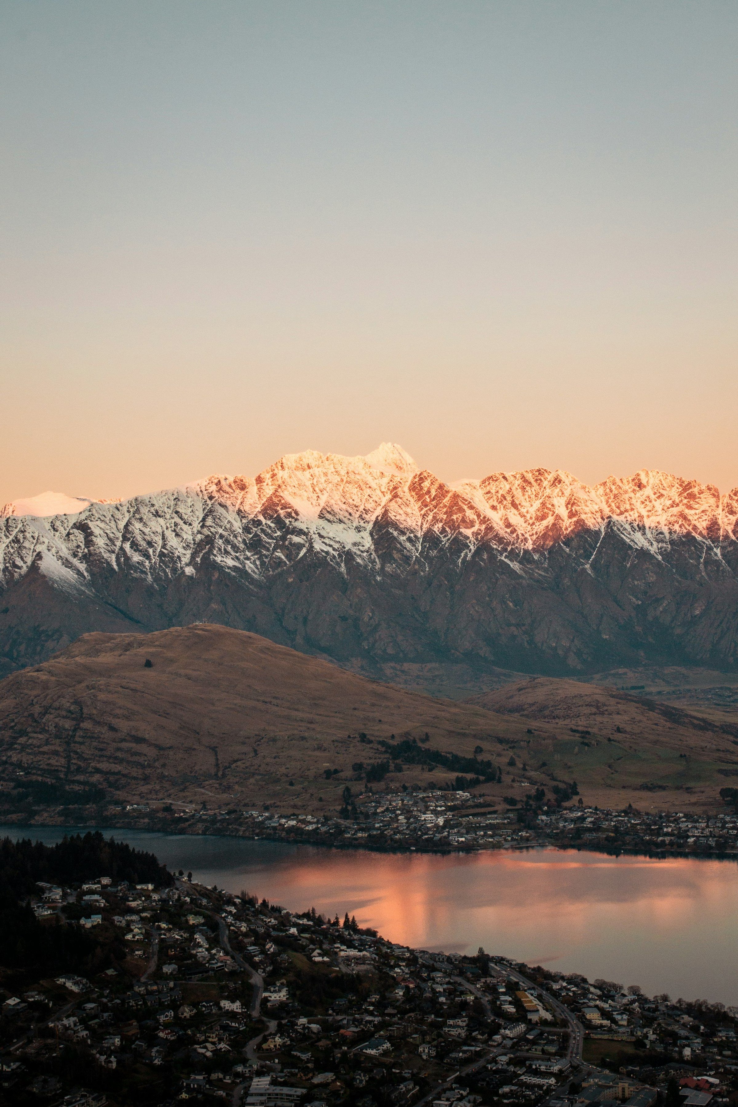 Panorama Queenstown musim dingin: pegunungan The Remarkables bersalju, Danau Wakatipu yang tenang, dan kota di tepiannya saat cahaya emas senja, Selandia Baru