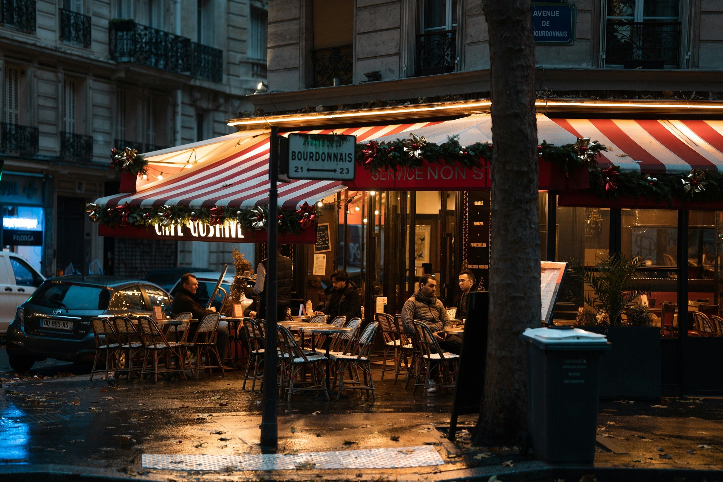 Teras kafe Paris di malam hari dengan lampu hangat dan pengunjung duduk di luar.