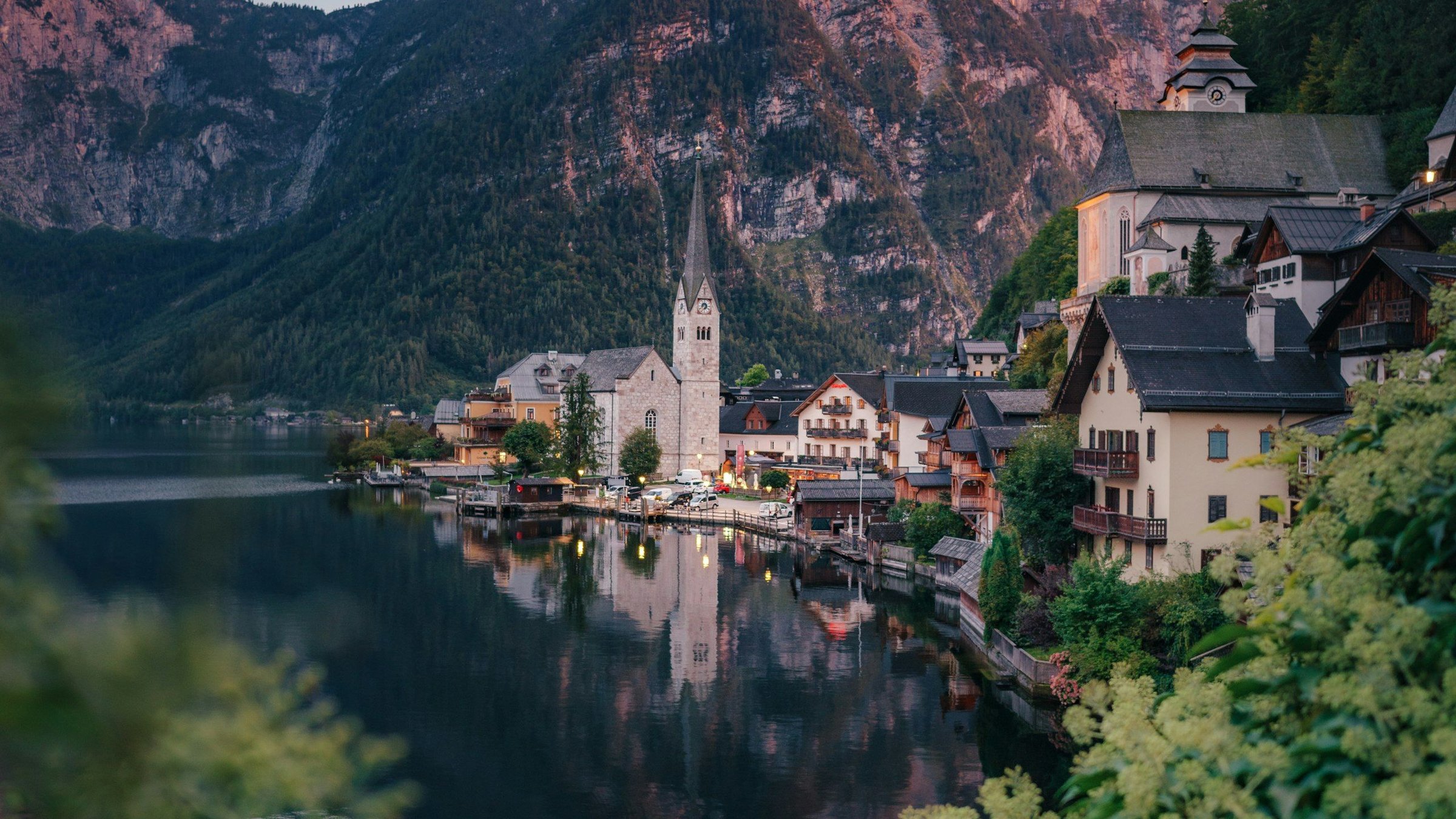 Desa Hallstatt di Austria dengan menara gereja, rumah kayu di tepi danau, refleksi air yang tenang, dan Pegunungan Alpen di belakang saat matahari terbenam