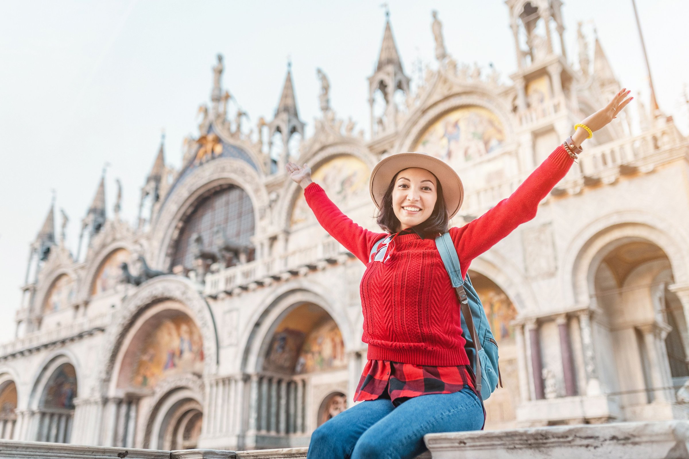Seorang wisatawan perempuan berbaju merah dan bertopi merentangkan tangan gembira di depan Basilika San Marco, Venice.