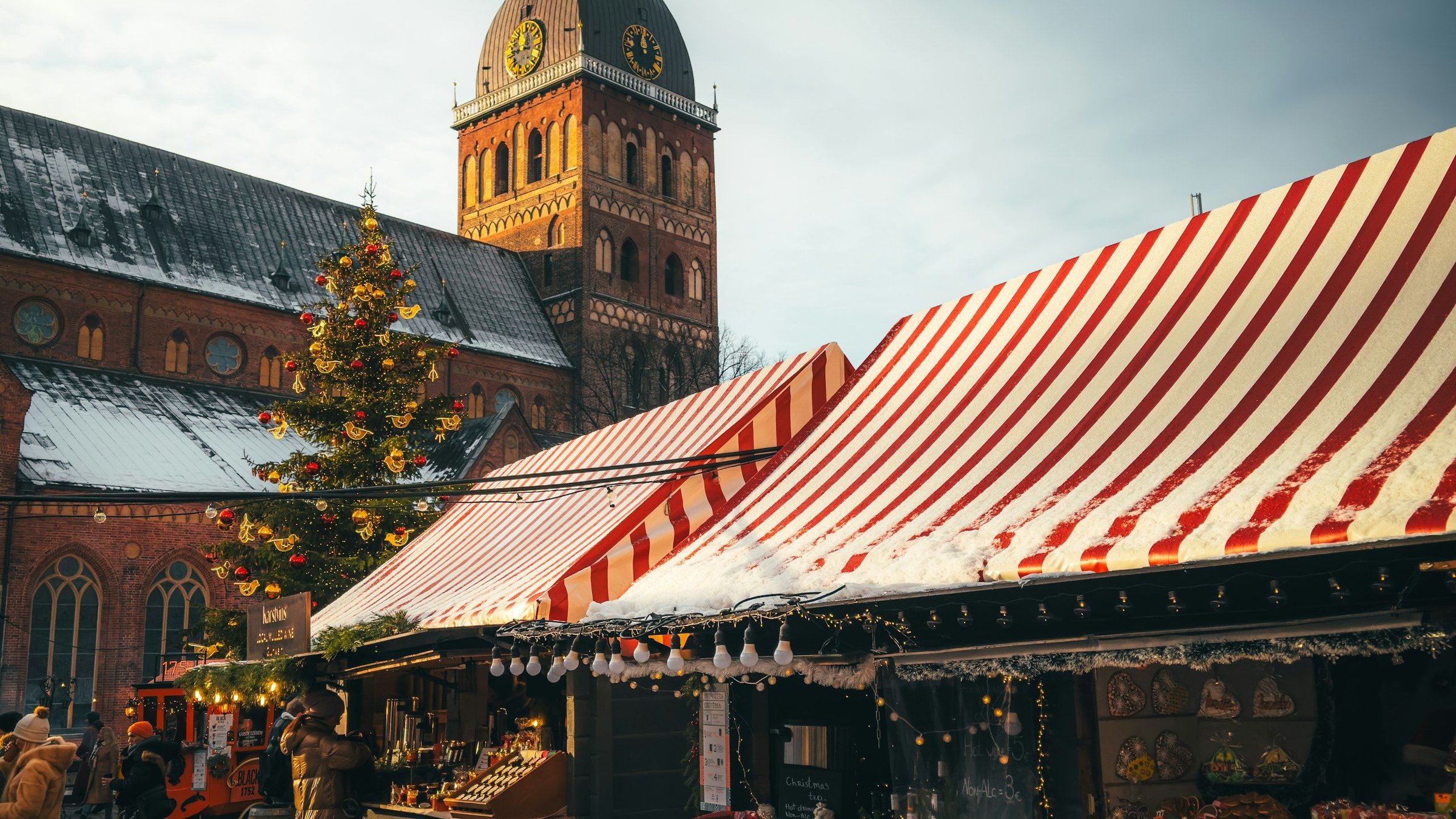 Pasar Natal Eropa di Riga dengan kios bergaris merah putih tertutup salju, pohon Natal berhias lampu, dan menara katedral berjam di latar saat musim dingin