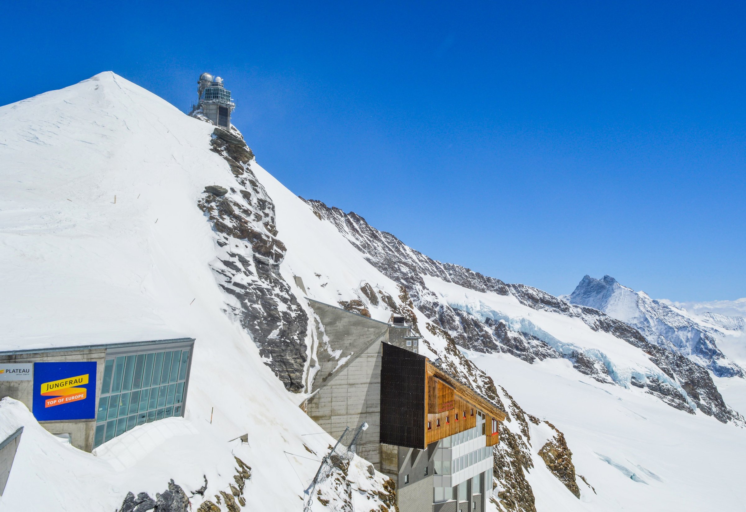Stasiun puncak Jungfraujoch bersalju dengan papan nama Jungfrau di bawah langit biru cerah