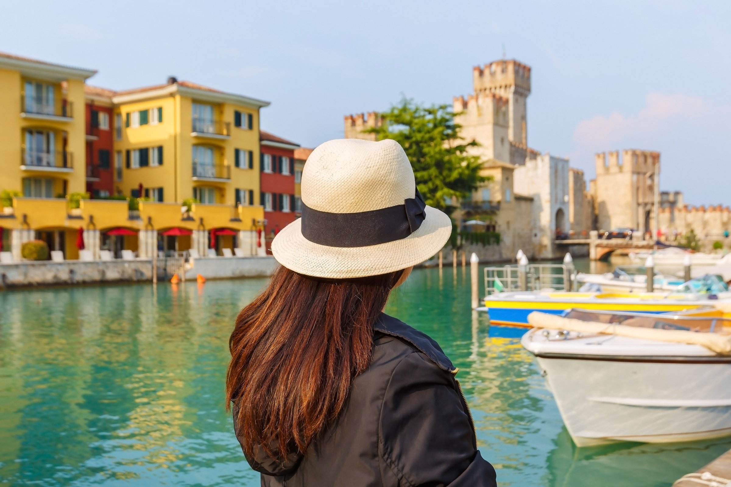 Seorang wisatawan perempuan bertopi memandang pelabuhan kecil dan Kastil Scaligero di Sirmione dengan rumah warna-warni.