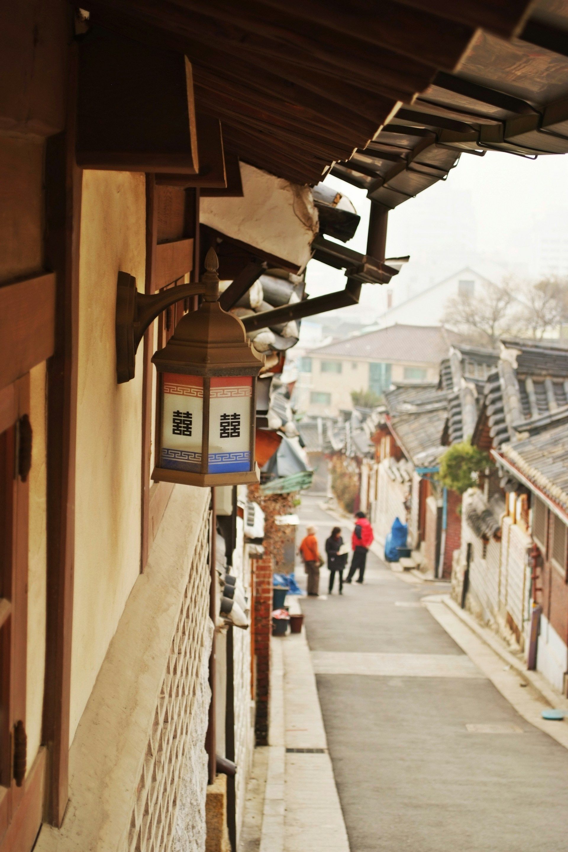 Gang sempit Bukchon Hanok Village di Seoul dengan lentera kayu motif double-happiness dan deretan atap genteng hanok melengkung
