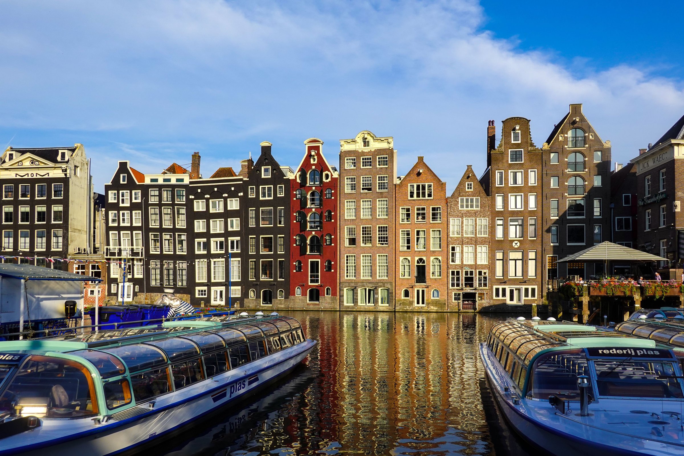 Deretan rumah kanal Amsterdam berwarna-warni dengan perahu wisata di bawah langit biru cerah