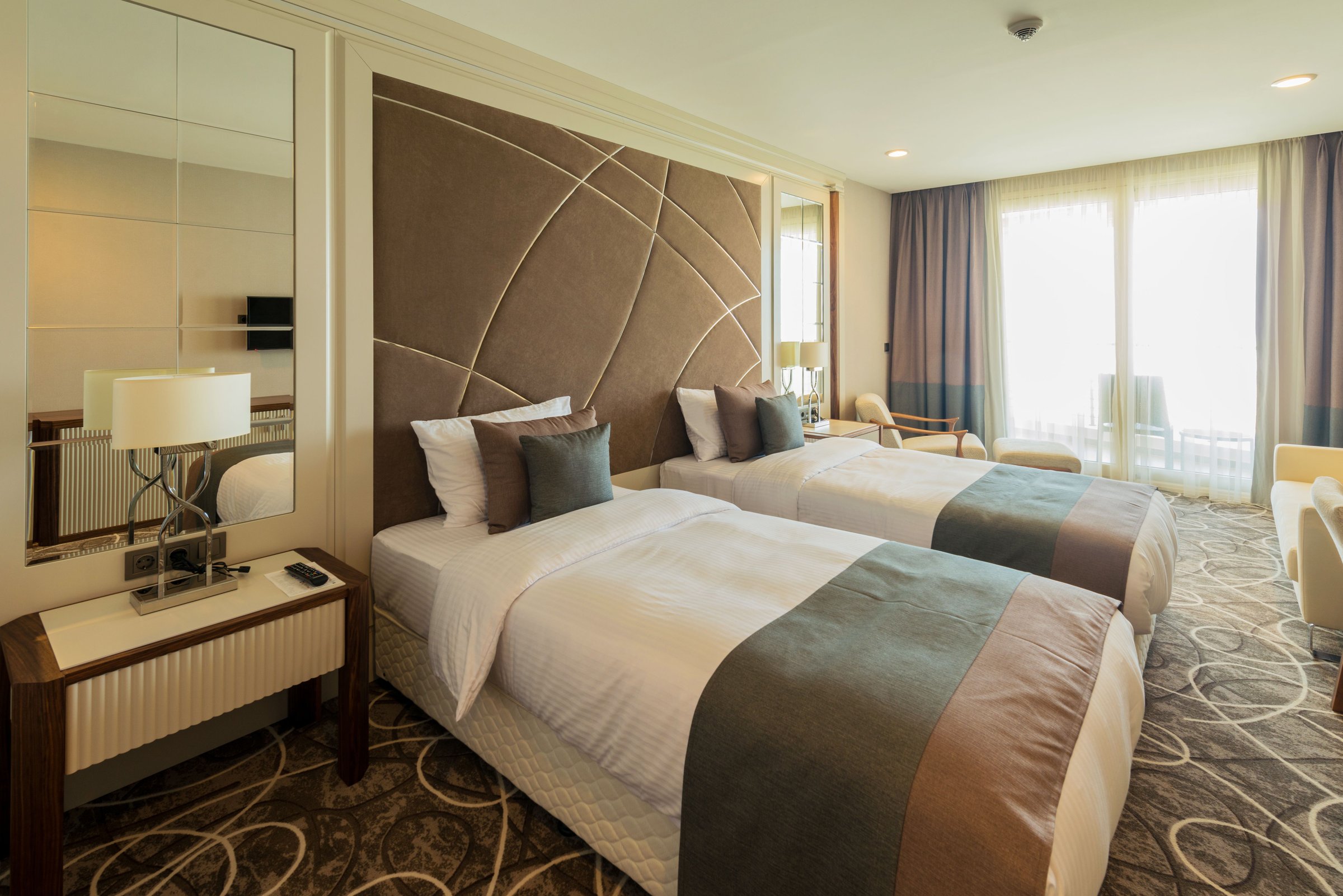 Kamar hotel dengan dua tempat tidur, headboard bermotif, dan jendela besar bertirai tipis