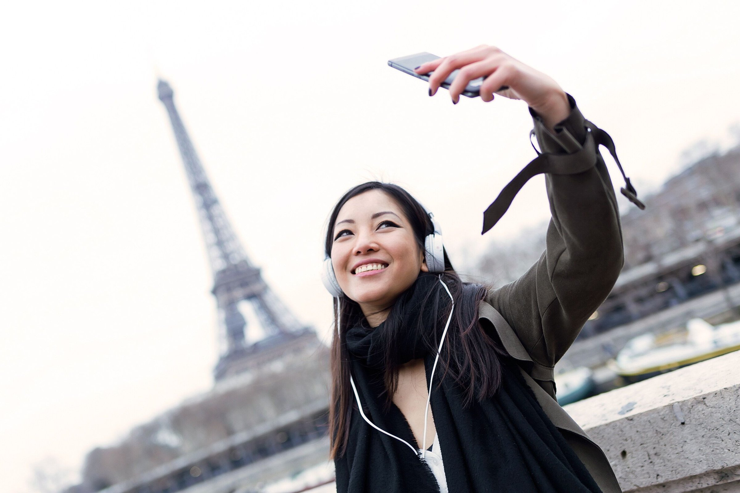 Perempuan Asia berjaket hangat dan berkalung syal tersenyum berswafoto dengan Menara Eiffel di belakangnya.