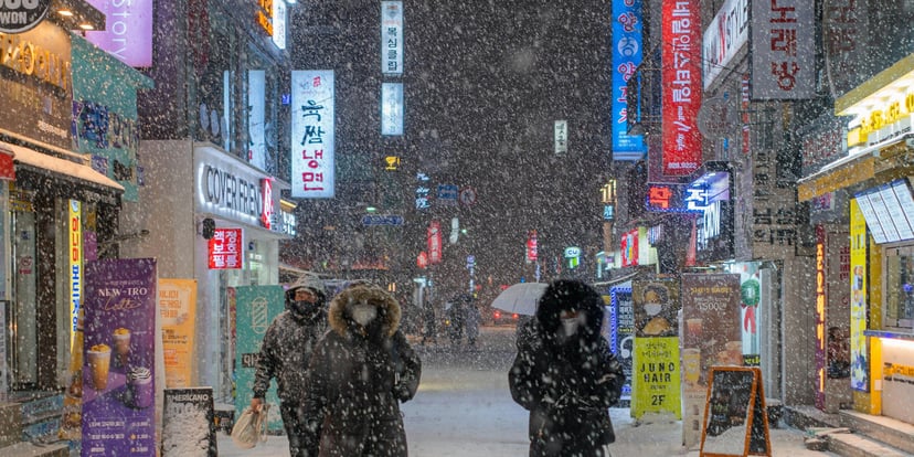 Jalanan Seoul diguyur salju malam hari, papan neon menyala dan orang berjalan menembus turunnya salju.