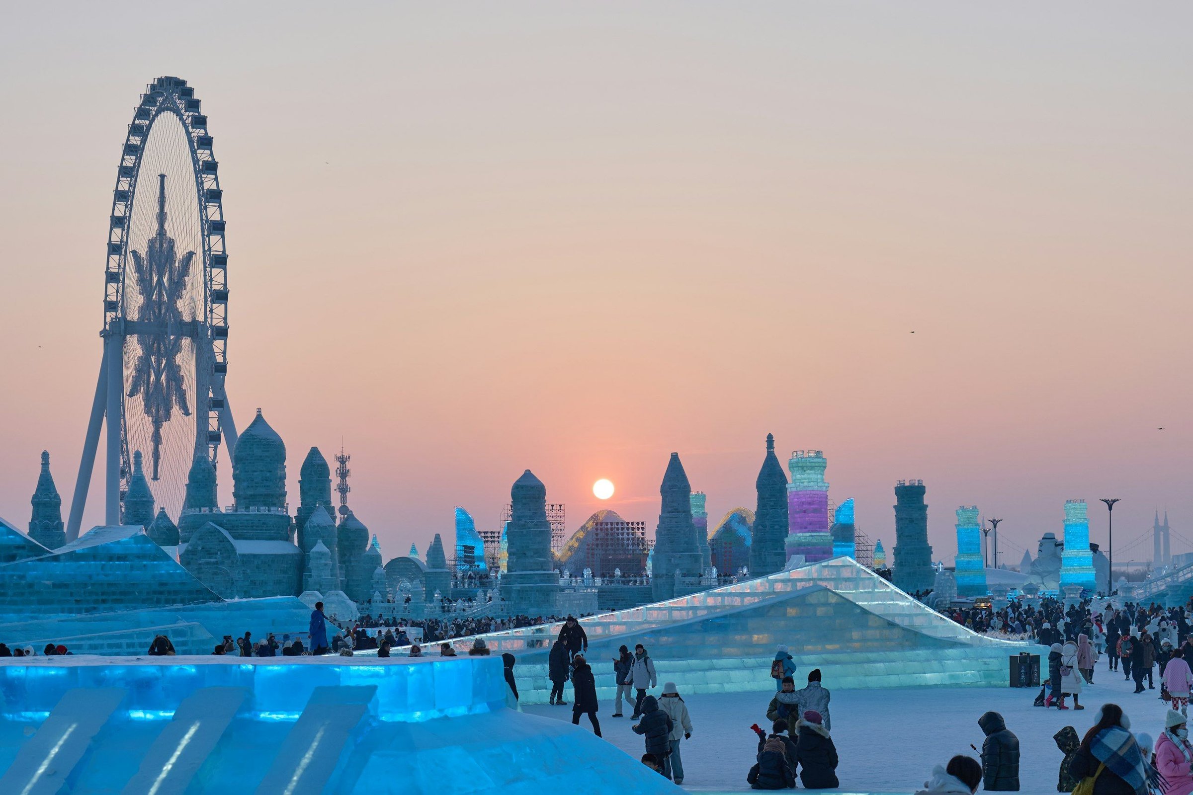 Harbin Ice and Snow World saat senja, dengan istana es raksasa yang menyala biru dan ungu, kincir ria, serta pengunjung di plaza bersalju, China