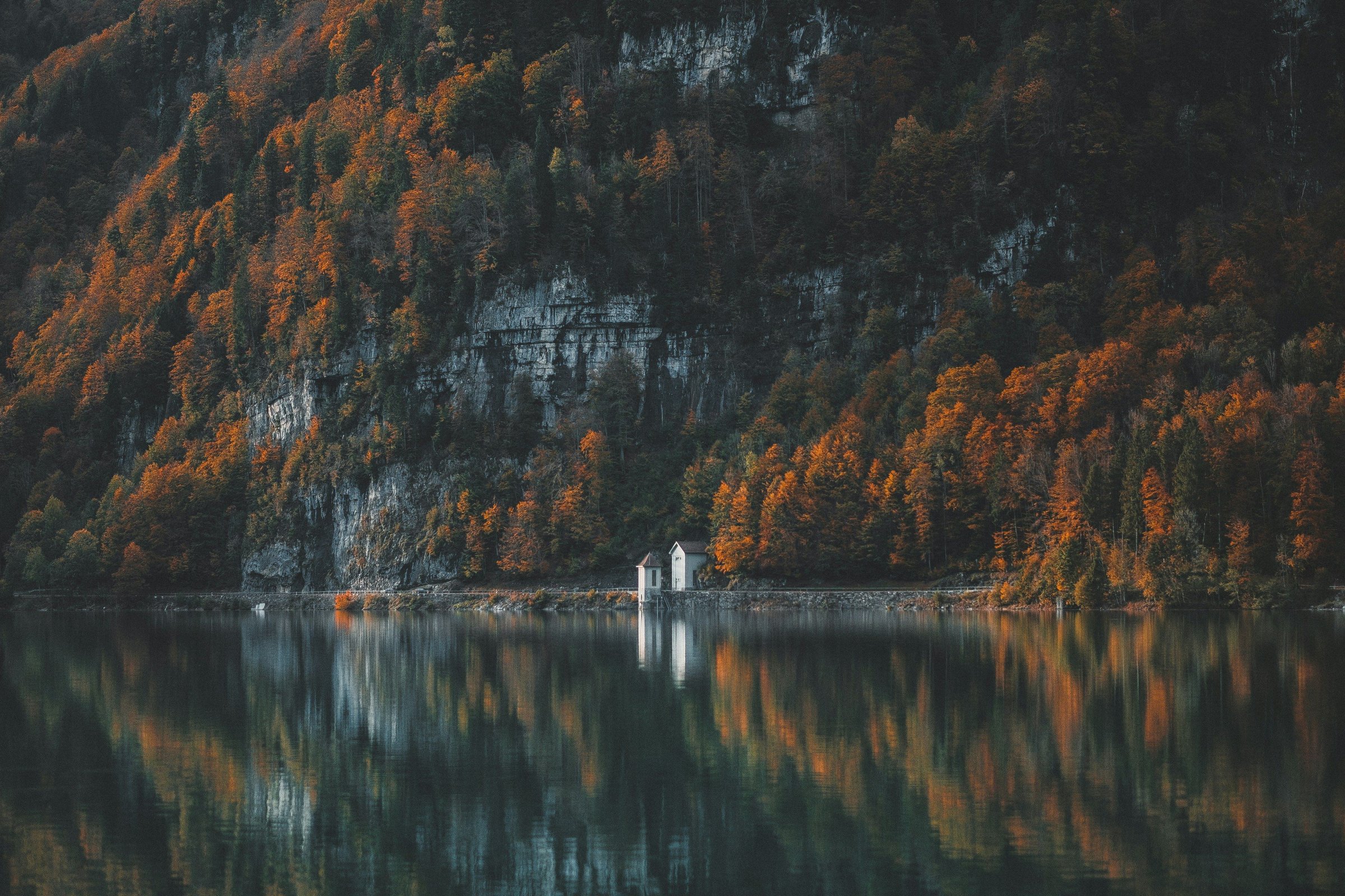 Danau alpine dengan lereng dedaunan musim gugur dan kabin kayu, refleksi tenang.
