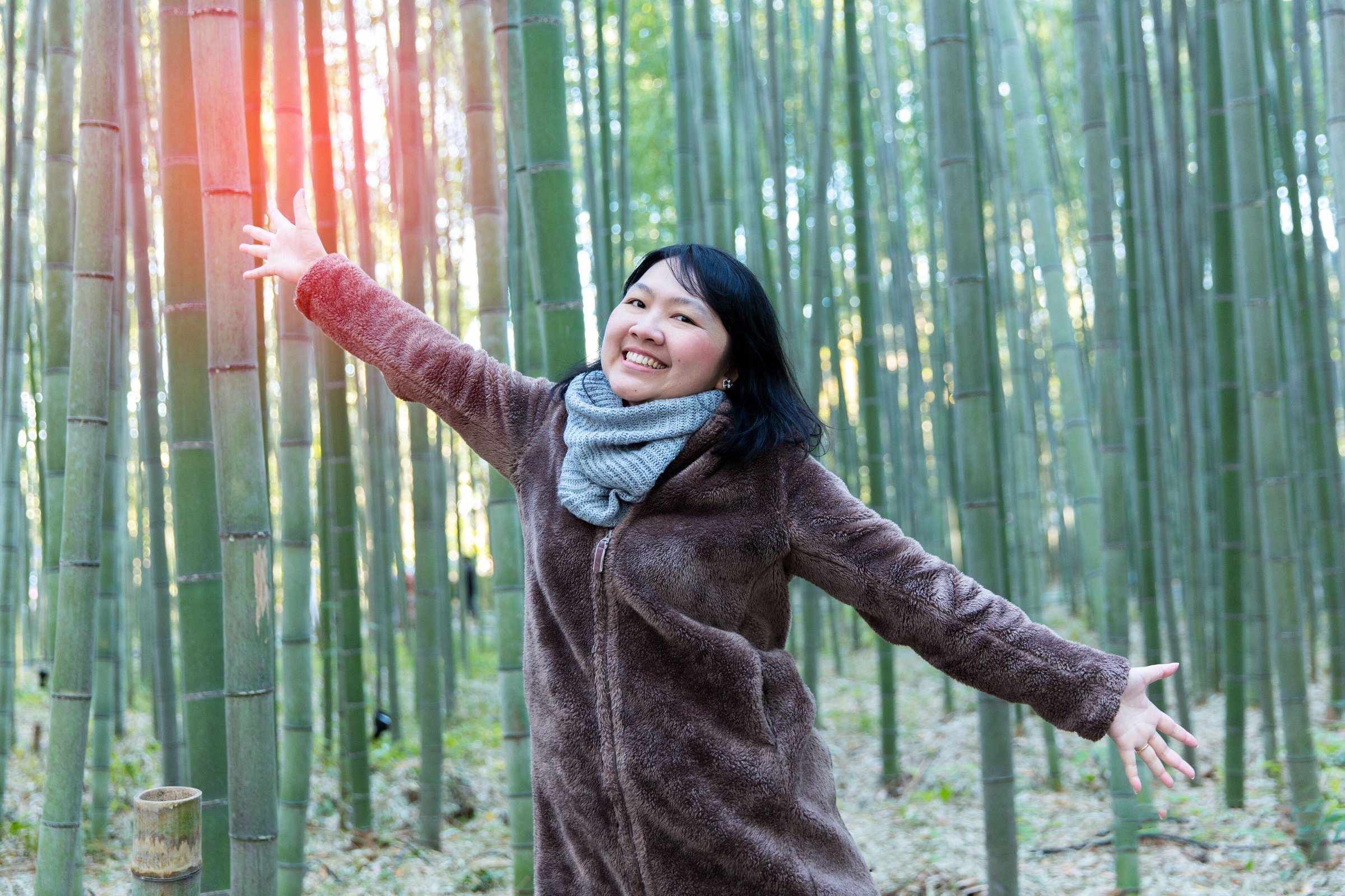 Perempuan berjaket bulu dan syal membentangkan tangan di hutan bambu Arashiyama, Kyoto