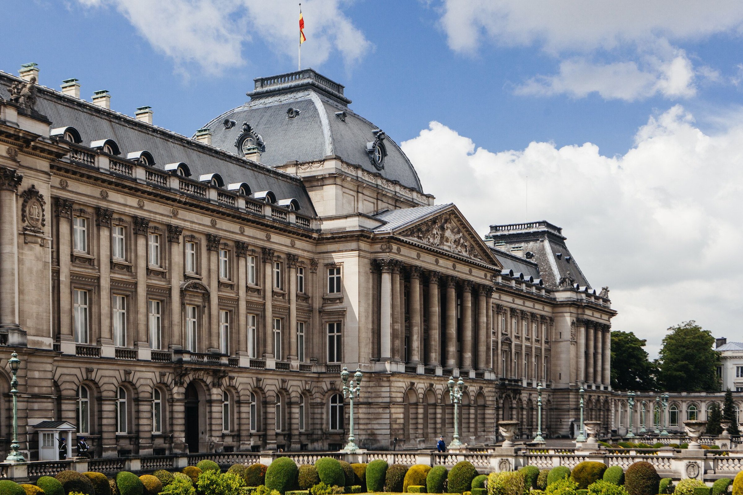 Royal Palace Brussels dengan taman pagar tanaman di depannya dan awan putih di langit biru.
