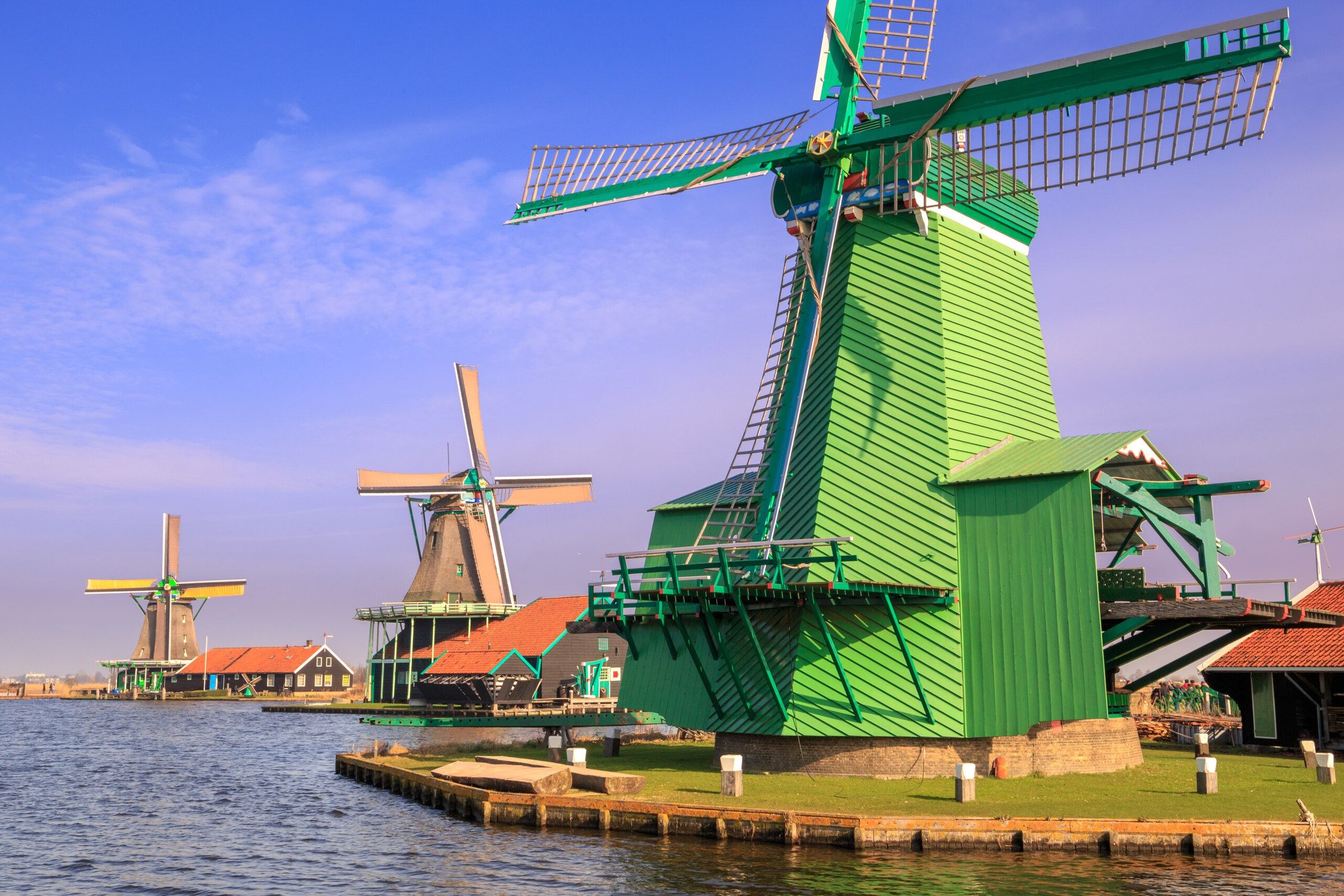 Kincir angin hijau dan merah di tepi sungai Zaanse Schans di hari cerah.