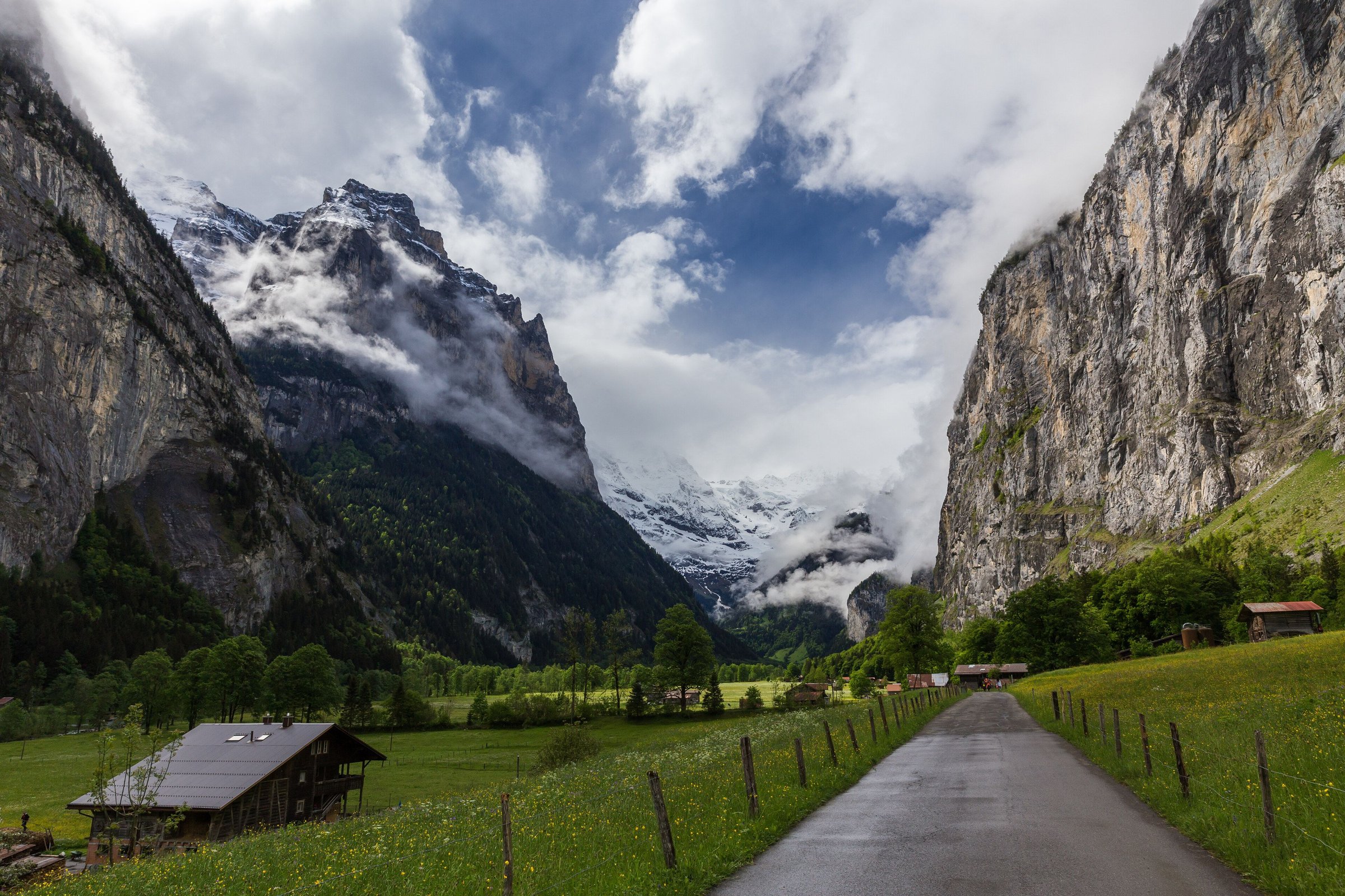 Jalan beraspal di lembah Lauterbrunnen menuju tebing dan puncak bersalju yang berselimut awan, rumput mulai hijau.
