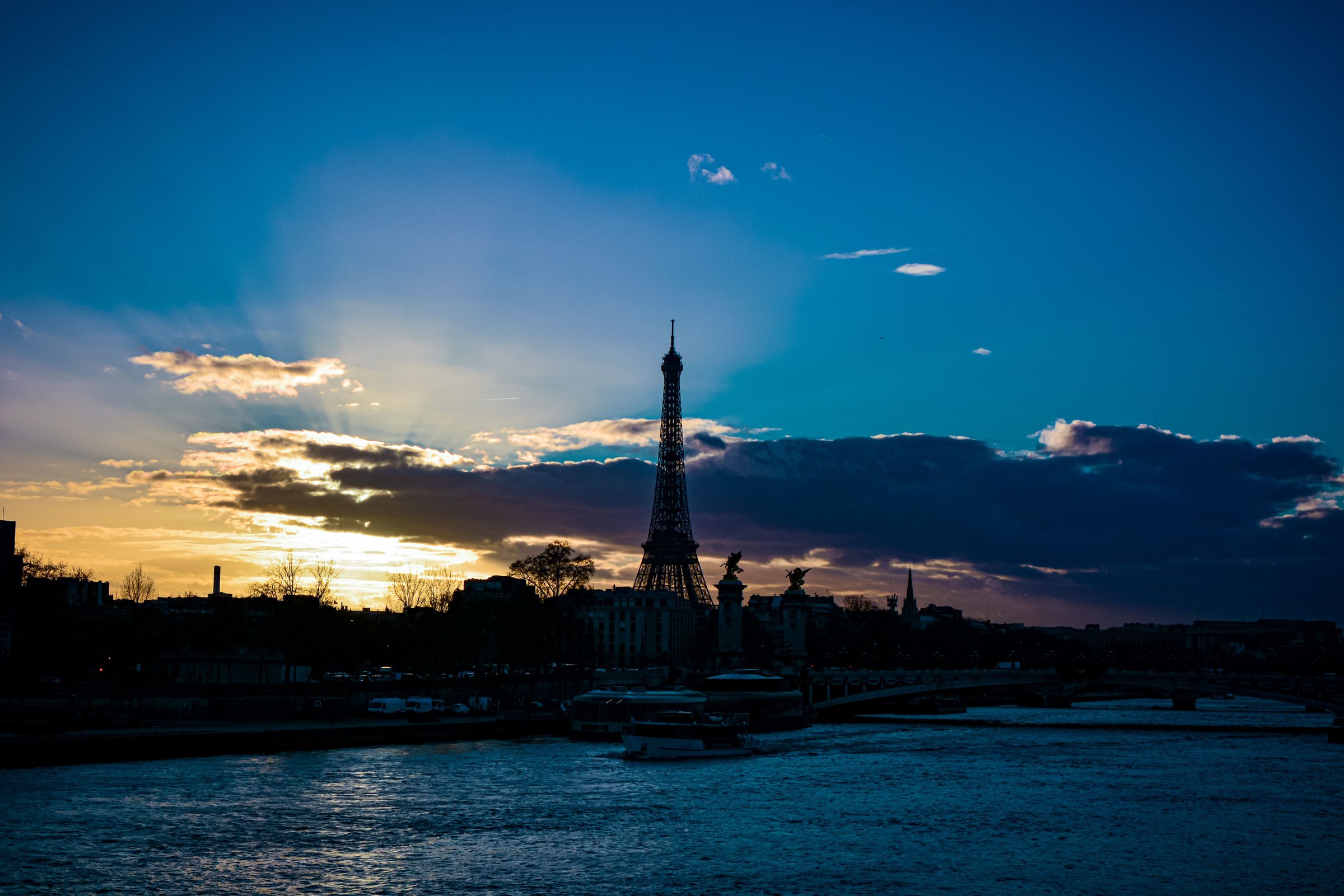 Siluet Menara Eiffel di tepi Sungai Seine saat senja dengan langit biru dramatis