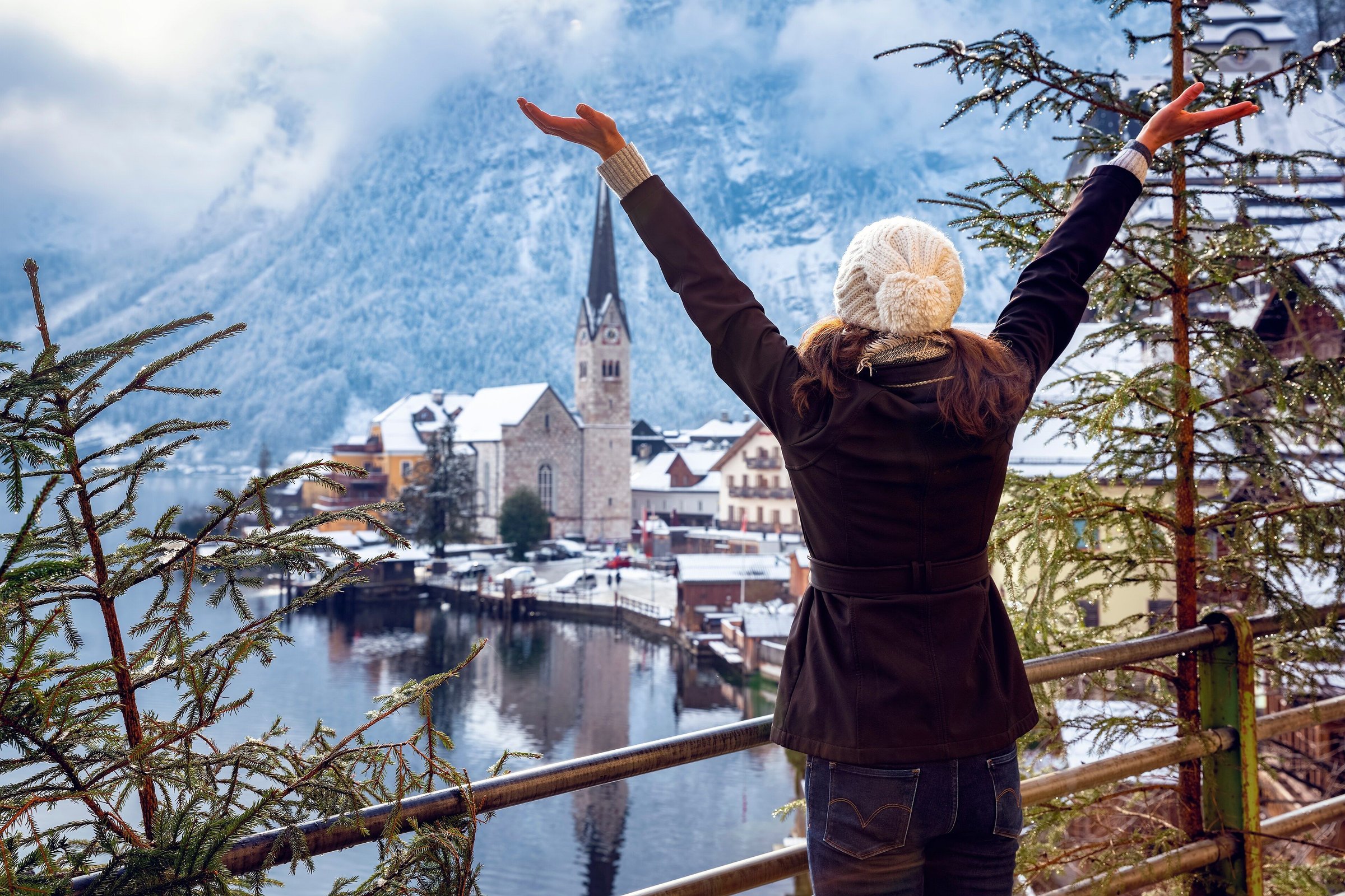 Wisatawan membentangkan tangan menghadap desa Hallstatt bersalju di tepi danau