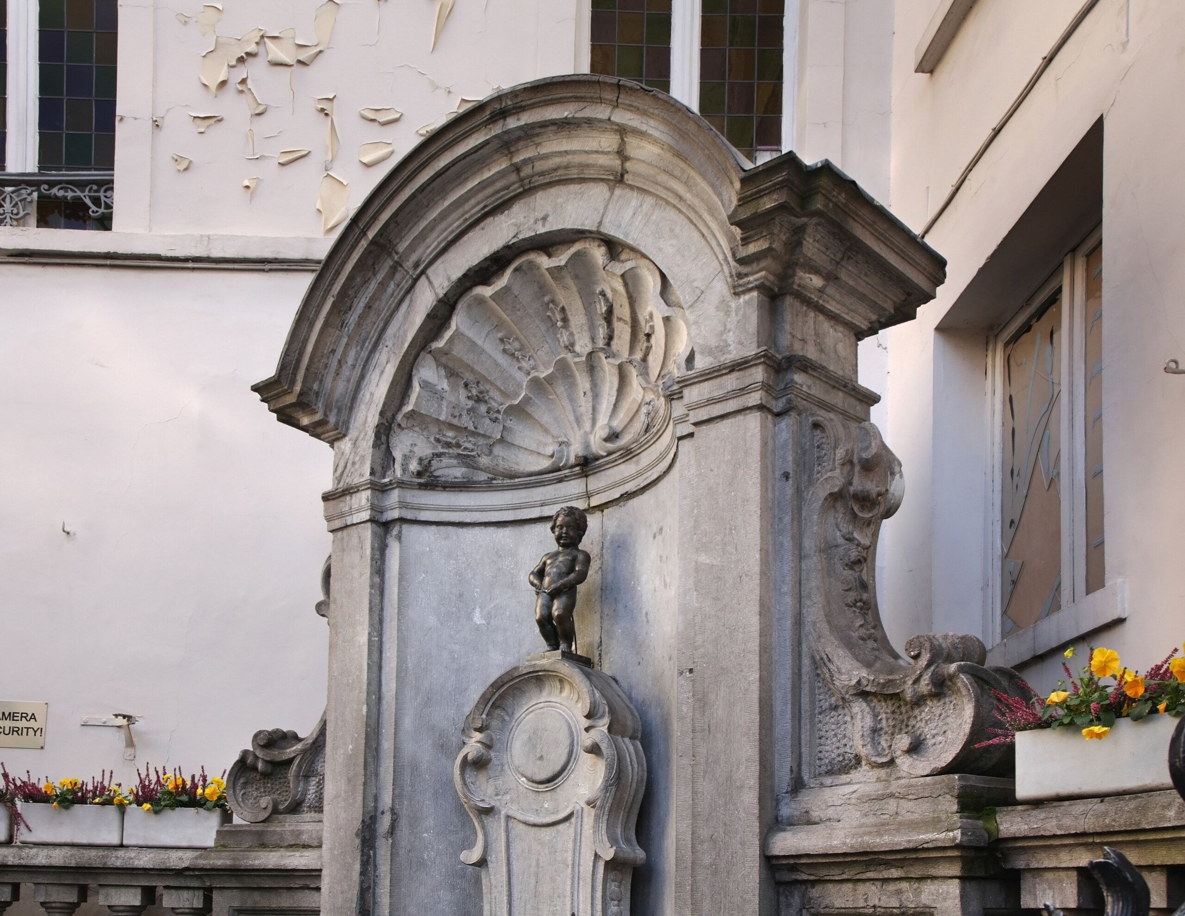 Patung Manneken Pis di relung batu dengan kotak bunga kuning di Brussels.