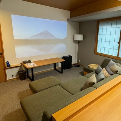 The Two Bedroom Theater Suite Living area has a large couch with throw pillows and ottoman footrests. There Japanese-style paper blinds on the windows and a projector for watching TV or movies on the wall.
