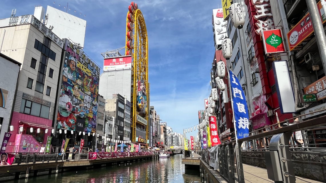 Daytime view of Dotonbori, Osaka, with colorful billboards, a ferris wheel, and vibrant banners along the canal.