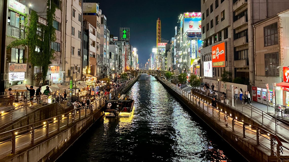 Night view of a lively canal in Dotonbori, Osaka, with illuminated buildings, bustling restaurants, and a boat on the water.