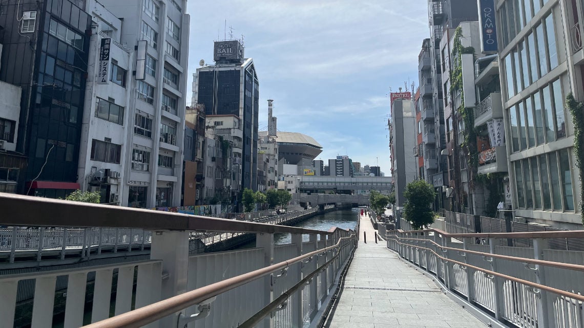 Dotonbori West Access Ramp - Daytime view of a quiet canal in Osaka, flanked by modern buildings, with a footbridge leading towards the city center.