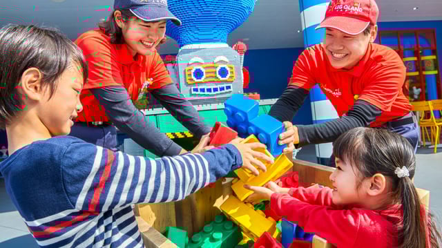 Children and staff build with large LEGO bricks at Legoland Japan, smiling and having fun near a LEGO robot figure.