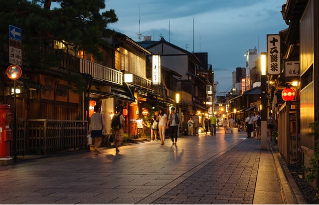 A lively, lantern-lit street in Japan at dusk, with traditional wooden buildings and pedestrians strolling along the cobblestone path.