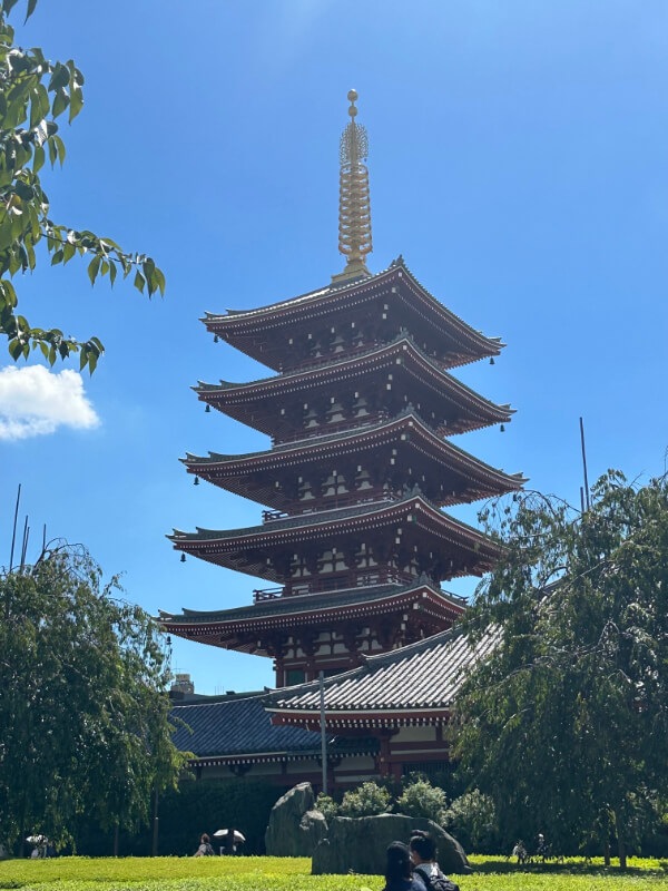 A towering five-story pagoda at Sensoji Temple in Tokyo stands against a bright blue sky. The pagoda’s traditional architecture is surrounded by lush greenery.