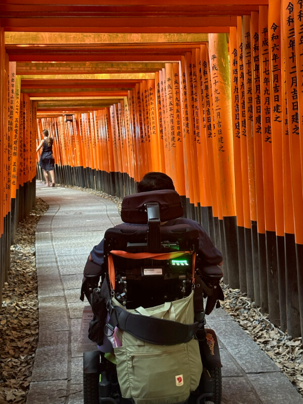Tayjus Surampudi navigates through the famous torii gates of Fushimi Inari Taisha Shrine in his power wheelchair. The vibrant orange gates create a tunnel-like path.