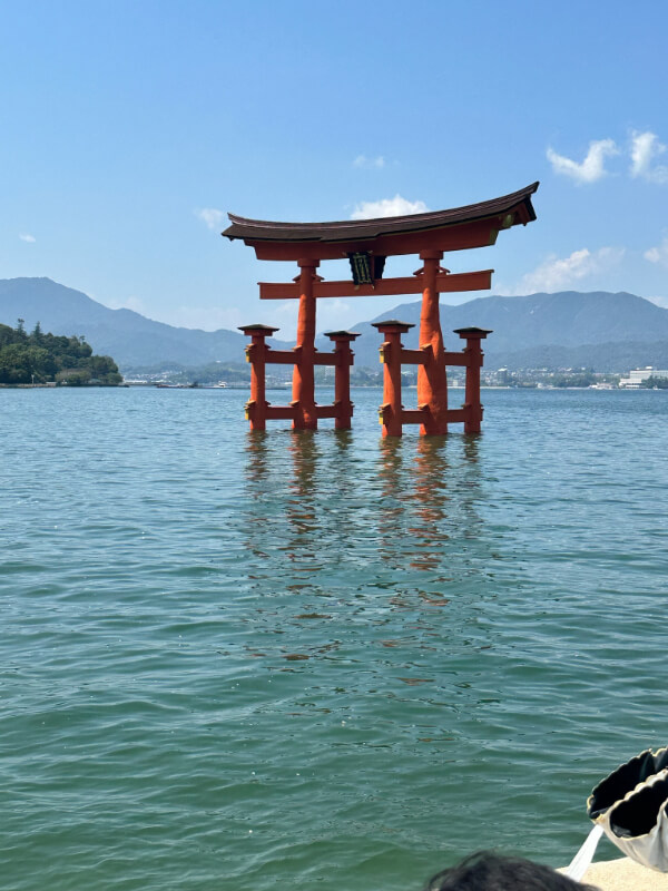 The iconic floating torii gate of Itsukushima Shrine rises from the calm waters of Miyajima Island. The bright orange gate contrasts with the blue sky and distant mountains.