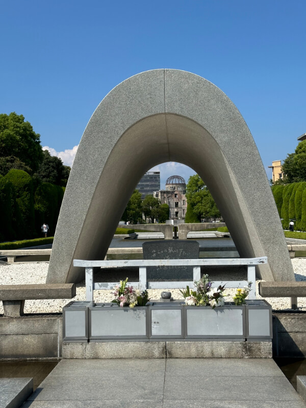 The Hiroshima Peace Memorial Cenotaph frames the Atomic Bomb Dome in the background. Flowers are placed at the memorial, symbolizing hope and remembrance under a clear blue sky.