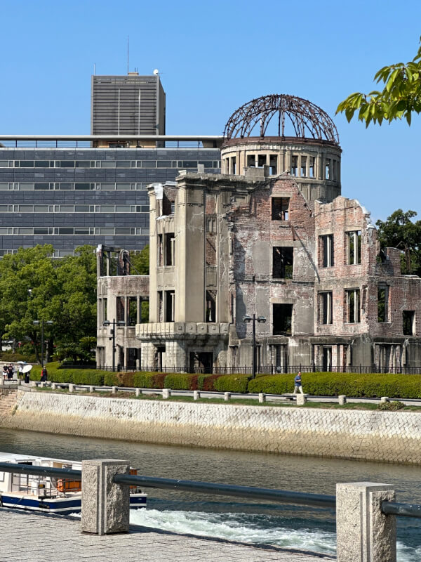The Atomic Bomb Dome in Hiroshima, a preserved ruin from the 1945 bombing, stands as a solemn reminder of history. The dome is set against a bright sky and a peaceful riverside scene.
