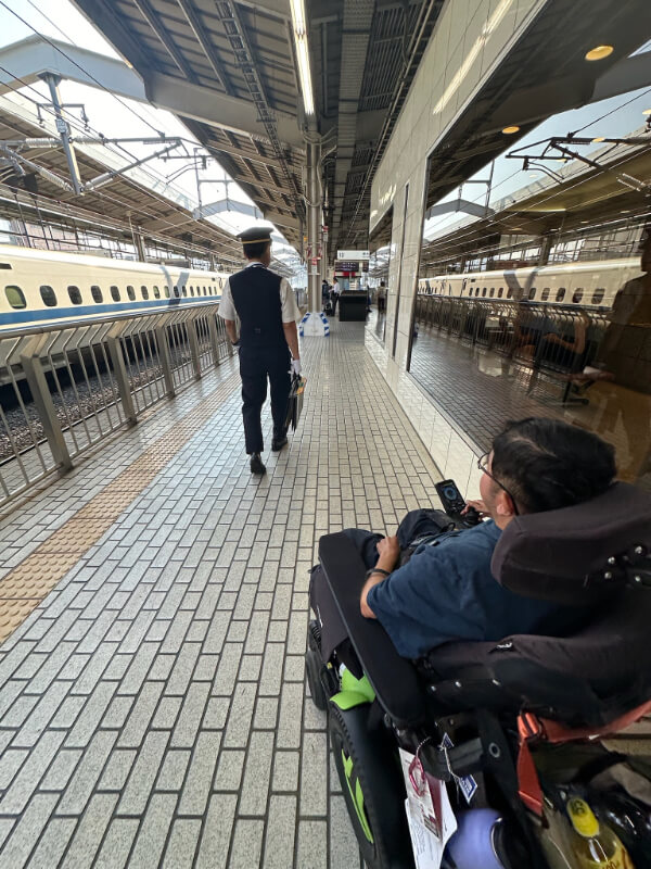 Tayjus Surampudi follows a station staff member on a Shinkansen platform, preparing to board the high-speed train. His power wheelchair is ready for a smooth travel experience in Japan.