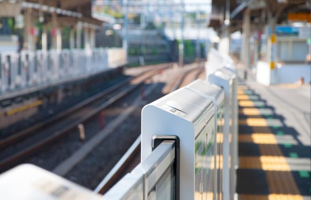 An empty train station platform with safety gates closed along the tracks. No train is visible, and sunlight illuminates the scene.