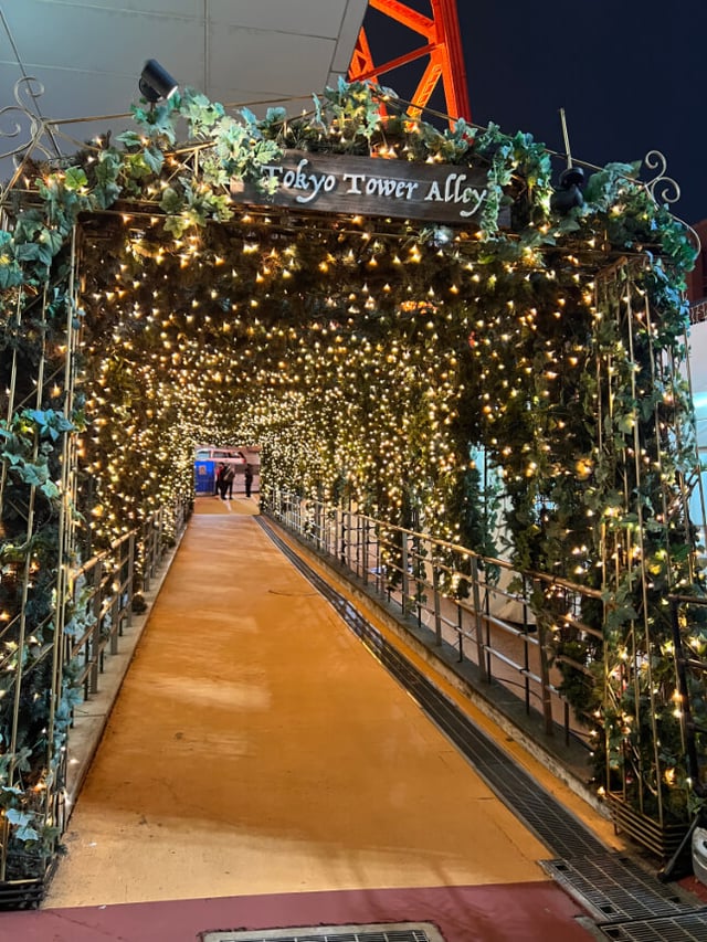 An illuminated tunnel labeled "Tokyo Tower Alley" decorated with greenery and string lights, leading to a festive walkway at night.