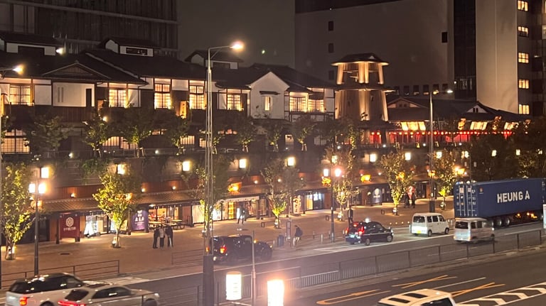 Night view of Toyosu Senkyaku Banrai, with a traditional two-story building lit by warm lights and lanterns. Illuminated storefronts and trees line the walkway.