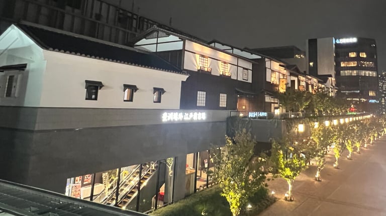 A night view of Toyosu Senkyaku Banrai showcasing a mix of white and dark wooden traditional-style architecture. Softly lit windows and glowing signage adorn the building. Below, a lit pathway lined with trees leads to storefronts.