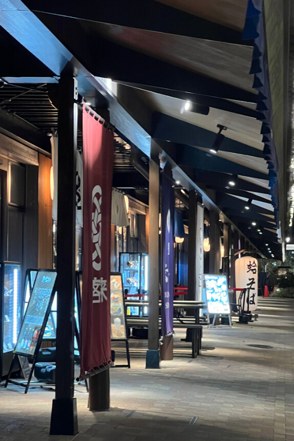 A night view of a covered walkway at Toyosu Senkyaku Banrai, lined with illuminated shop displays, colorful hanging banners with Japanese text, and wooden beams. Menu boards and signs showcase food offerings.