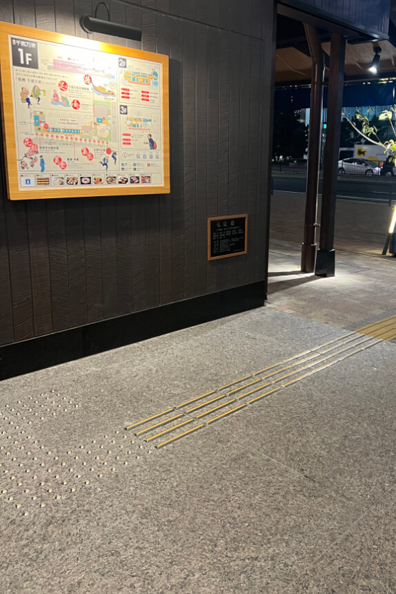 An accessibility-friendly walkway at Toyosu Senkyaku Banrai with tactile paving for visually impaired visitors. A brightly lit map of the building layout is mounted on a dark wooden wall.