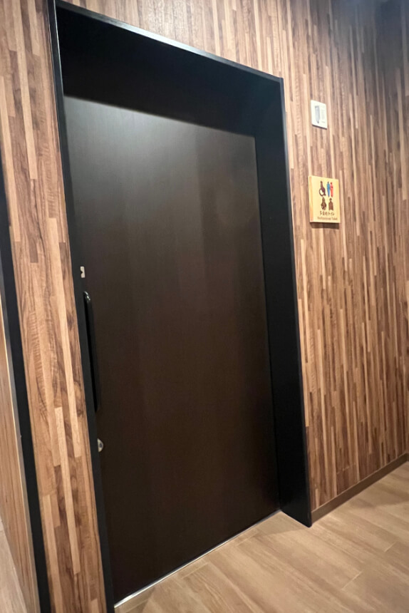 A dark wooden door to a multipurpose toilet, set within walls featuring a wood-pattern design. A sign next to the door displays icons indicating accessibility for wheelchair users, caregivers with children, and both male and female users.
