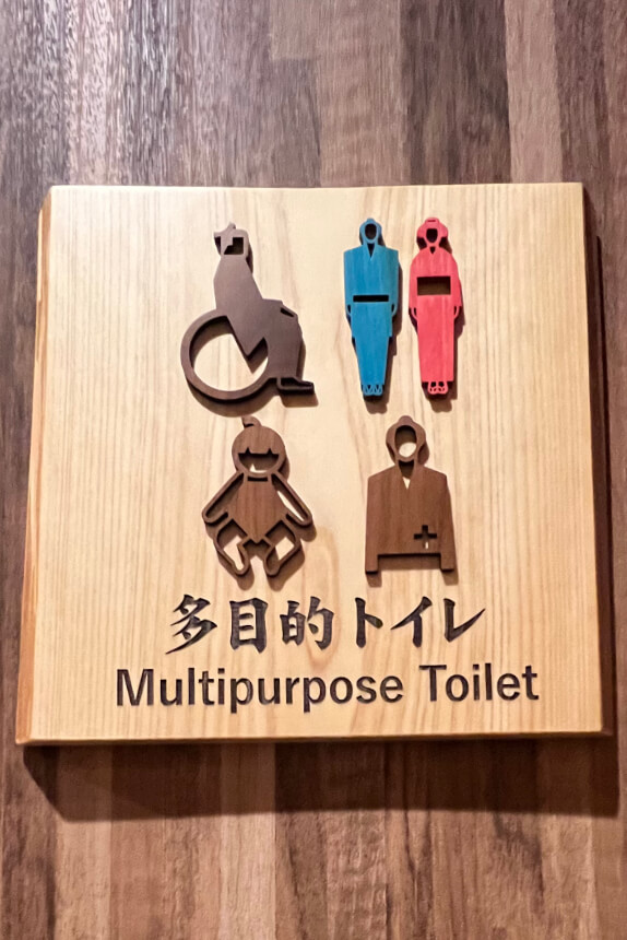 A wooden sign for a multipurpose toilet featuring icons for accessibility: a wheelchair, a caregiver with a child, and figures for male and female users. Below the icons, text reads "Multipurpose Toilet" in English and Japanese.