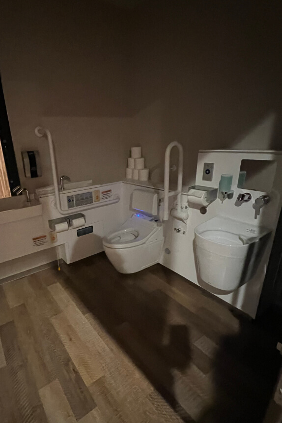 A spacious multipurpose restroom with a bidet toilet, safety handrails, and a wall-mounted sink for ostomate users.