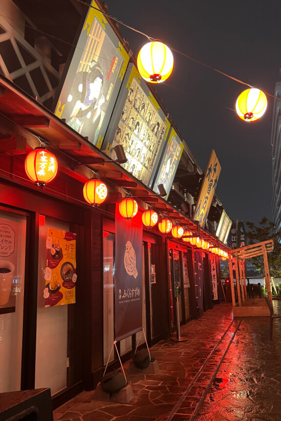 A vibrant night scene at Toyosu Senkyaku Banrai, featuring a row of shops adorned with red lanterns casting a warm glow. Illuminated traditional-style artwork and signs hang above the storefronts, with banners advertising food.