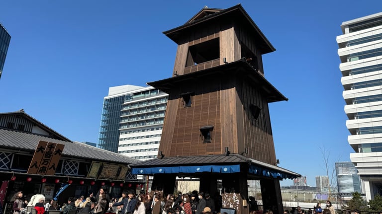 A tall wooden bell tower at Toyosu Senkyaku Banrai stands prominently under a bright blue sky. Crowds gather around its base.