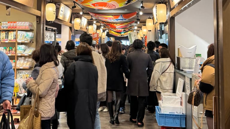A bustling and crowded indoor market at Toyosu Senkyaku Banrai, with shoppers filling the narrow walkway. Stalls on both sides display packaged goods and local items. Lanterns and colorful fish-themed banners hang overhead.