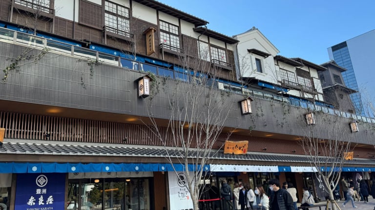 A traditional-style two-story building at Toyosu Senkyaku Banrai in Tokyo, featuring wooden facades, tiled roofs, and paper lanterns with Japanese text. The ground floor has blue fabric awnings with logos, shops, and people walking or standing in line. A traditional-style two-story building at Toyosu Senkyaku Banrai in Tokyo, featuring wooden facades, tiled roofs, and paper lanterns with Japanese text. The ground floor has blue fabric awnings with logos, shops, and people walking or standing in line.